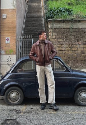 Chaqueta bomber de cuero marrón, jersey de cuello alto beige, pantalones blancos, zapatos negros, de pie al lado de un coche vintage azul marino en una calle.