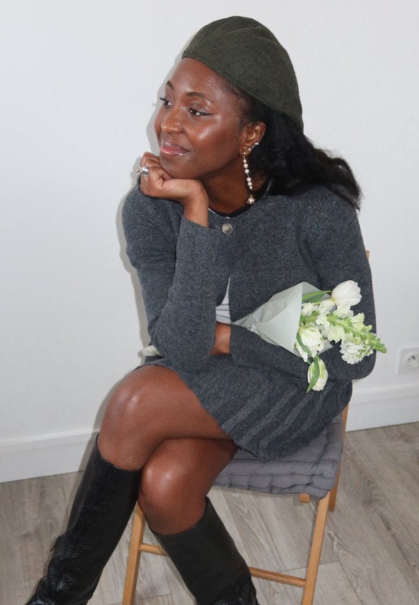 Femme en robe grise et chapeau vert assise sur une chaise, tenant des fleurs blanches, reposant le menton sur la main, souriant doucement sur le côté.