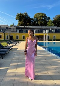 Abito lungo in satin rosa con scollatura a cowl, vestibilità slim e spalline sottili. Bracciale e borsetta in colori a contrasto. Ambientazione a bordo piscina.