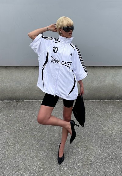 Chaqueta blanca oversize con rayas negras y el texto "TEAM GEIST", combinada con shorts de ciclismo negros y tacones de punta negros. Gafas de sol negras y bolso.