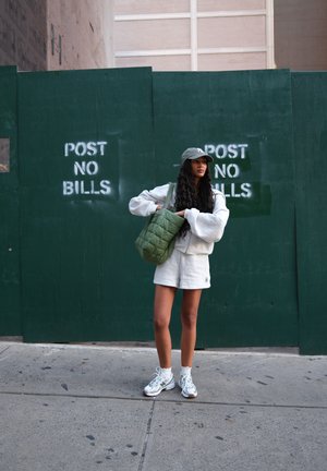 Mujer con atuendo blanco casual y gorra de pie en la acera, sosteniendo un bolso acolchado verde, frente a una pared verde con letreros de "NO ADJUNTAR FACTURAS".