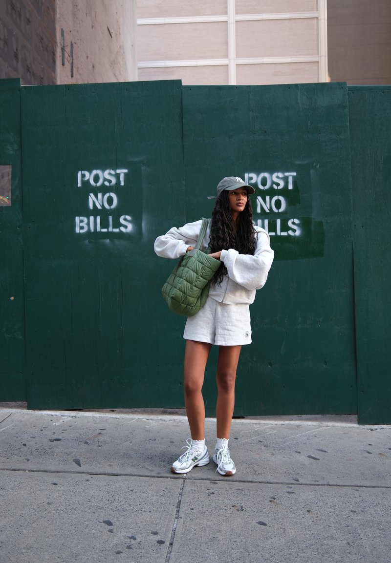 Una donna in un abbigliamento casual bianco e cappellino si trova sul marciapiede, mentre tiene una borsa trapuntata verde, di fronte a un muro verde con cartelli che dicono "POST NO BILLS".