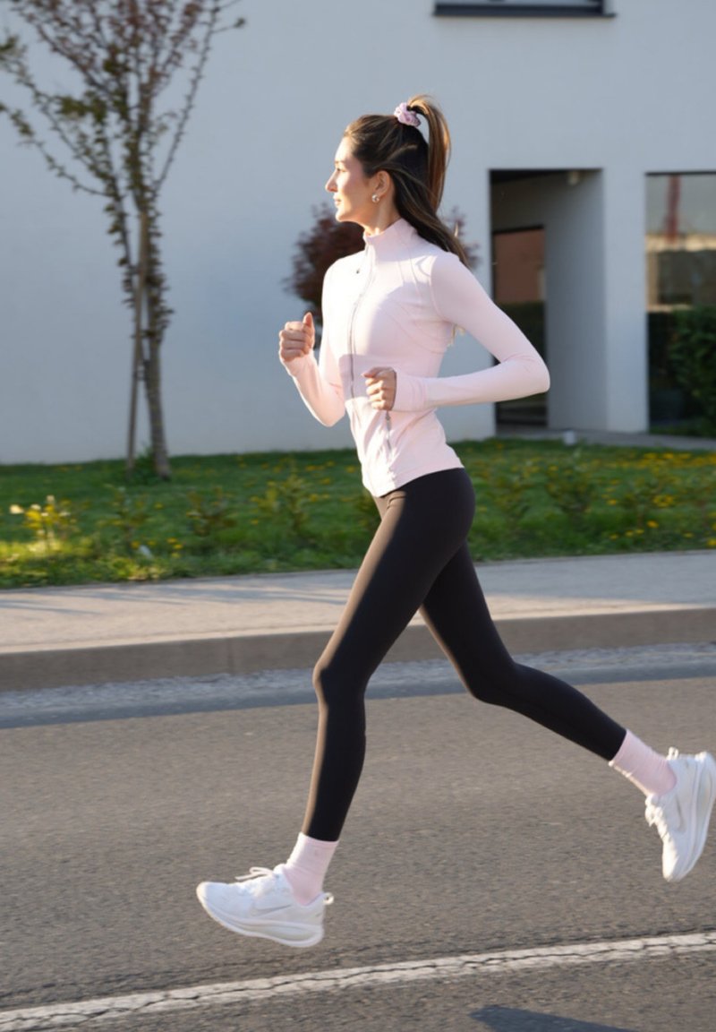 Eine Frau joggt im Freien an einem sonnigen Tag, trägt eine hellrosa Jacke, schwarze Leggings und weiße Turnschuhe. Im Hintergrund sind Gebäude und Grünflächen zu sehen.