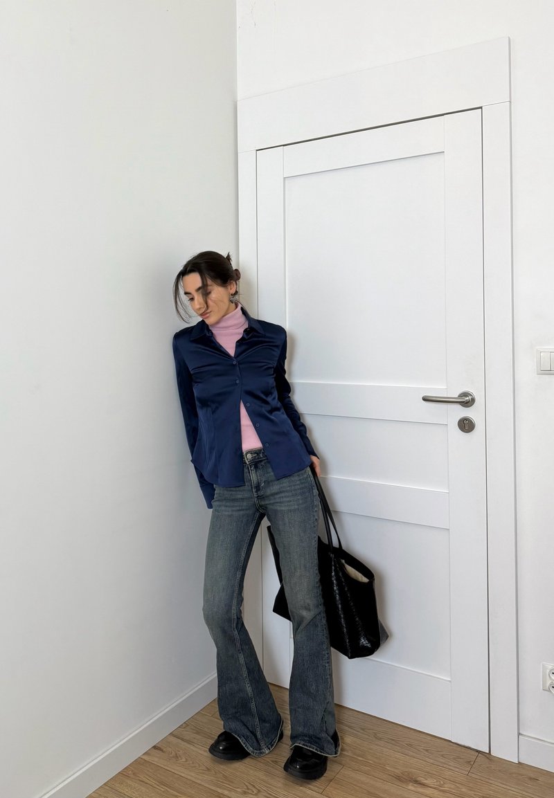 Navy satin button-up shirt over a light pink turtleneck, paired with flared denim jeans and black platform shoes, holding a black tote bag.