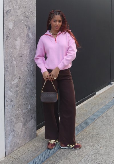 Sudadera rosa con detalle de cremallera, combinada con pantalones de pierna ancha marrones. Los accesorios incluyen un pequeño bolso oscuro y zapatillas multicolor.