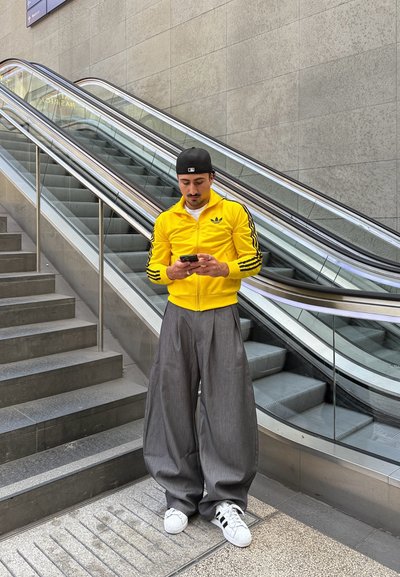 Chaqueta amarilla con cremallera y rayas negras, pantalones anchos grises y zapatillas blancas, de pie en unas escaleras cerca de una escalera mecánica, mirando un teléfono.