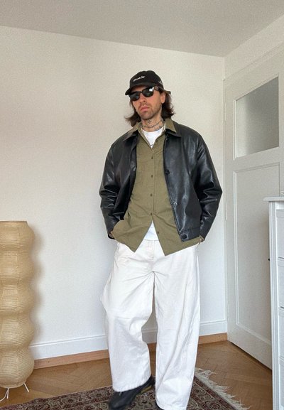 Hombre con gorra negra y gafas de sol, chaqueta de cuero negra, camisa verde oliva y pantalones anchos blancos, de pie en el interior cerca de una puerta y una lámpara.