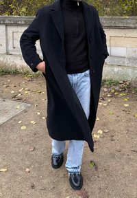 Μαύρο παλτό με γιακά, φορεμένο πάνω από μια ριγωτή μαύρη ζακέτα. Συνδυασμένο με ανοιχτό μπλε τζιν και μαύρα παπούτσια, σε ένα χαλίκι με φύλλα.