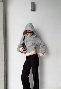 Sweat à capuche gris raccourci avec fermeture éclair et capuche, associé à un pantalon long noir. Accessoires compris : un petit sac noir et des lunettes de soleil.