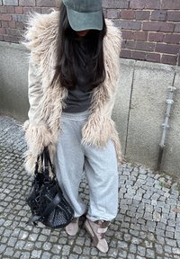 Femme debout sur des pavés, portant un manteau en fourrure beige, une chemise foncée, un pantalon de survêtement gris, des baskets beiges, une casquette verte et tenant un sac à main noir tressé.