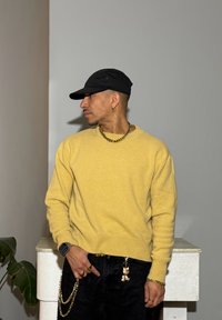 Žltý pletený sveter, čierna čiapka, zlatý retiazkový náhrdelník, tmavé džínsy s ozdobným retiazkou a príveskom, na neutrálnom pozadí.