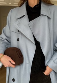 Femme portant un manteau bleu clair et un haut en dentelle noire, tenant une pochette en tissu marron pliée dans la main gauche, la main droite dans la poche.