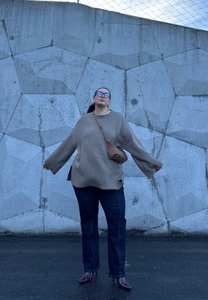 Suéter beige de gran tamaño con textura acanalada, jeans azul oscuro y bolso cruzado marrón. De pie frente a una pared de concreto texturizada.