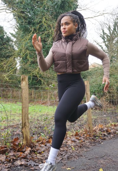 Chaleco de plumas marrón sobre una camiseta de manga larga beige, leggings negros de cintura alta, calcetines blancos y zapatillas de correr grises; corriendo al aire libre.