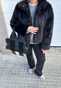 Personne portant une veste en fausse fourrure noire, un pull gris, un pantalon noir, des baskets argentées, tenant un sac à main en cuir noir avec des franges, debout sur un trottoir carrelé.