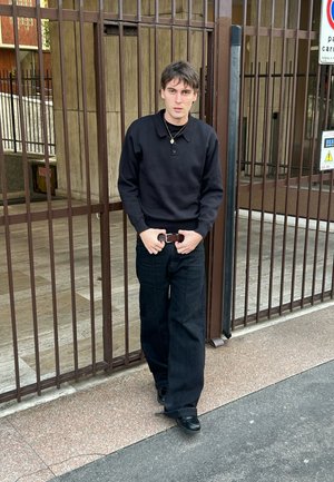 Joven con suéter negro y pantalones anchos negros está de pie frente a una puerta de metal marrón en la acera de una ciudad.