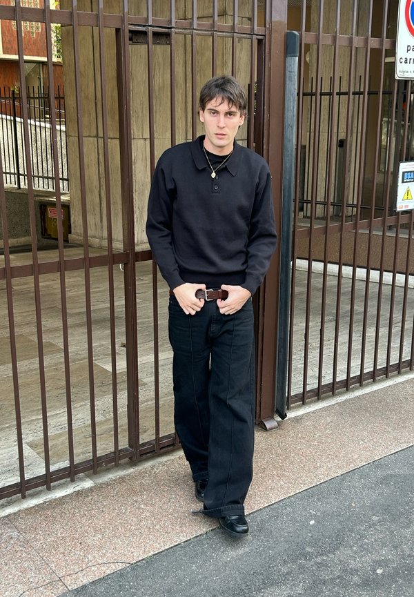 Jeune homme en pull noir et pantalon large noir se tenant devant une porte en métal marron sur un trottoir en ville.