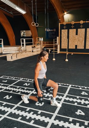 Mujer en el gimnasio haciendo zancadas con kettlebells sobre una esterilla numerada en blanco y negro bajo arcos de madera y anillas gimnásticas.