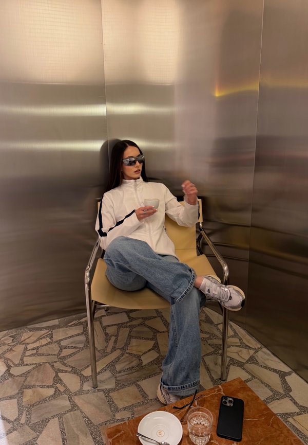 Femme portant des lunettes de soleil et une veste blanche, assise en tailleur sur une chaise, tenant une tasse, avec un téléphone et un verre d'eau sur une table proche.