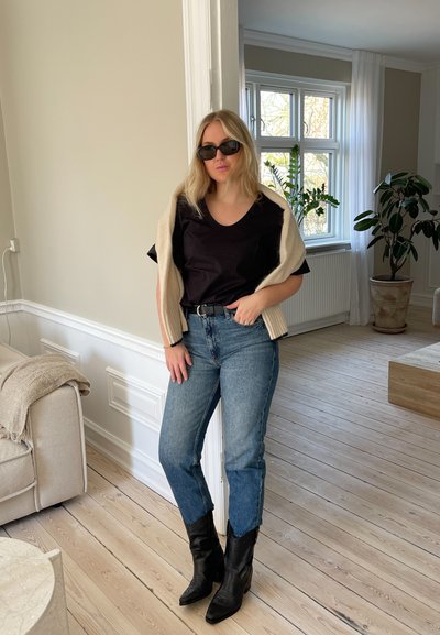 Camiseta negra, suéter crema casualmente colgado, jeans azules y botas negras de tobillo. Habitación con suelo de madera y plantas de interior visibles.
