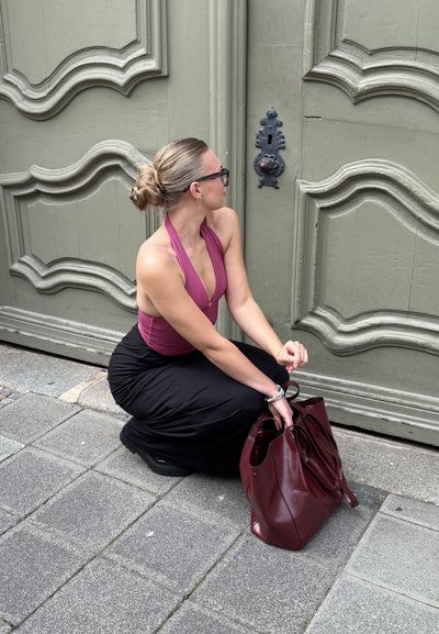 Una persona se agacha cerca de una puerta verde, vistiendo un top halter rosa profundo y una falda negra, sosteniendo un bolso de cuero color burdeos.