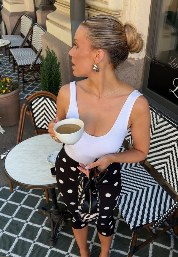 Femme avec un chignon blond tenant une tasse de café et un sac à main, debout à côté d'une table de café extérieure et de chaises à motifs noir et blanc.