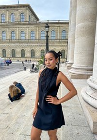 Jonge vrouw met gevlochten haar in een zwarte mouwloze jurk poseert op de stenen treden van een historisch gebouw met grote kolommen en gebogen ramen.