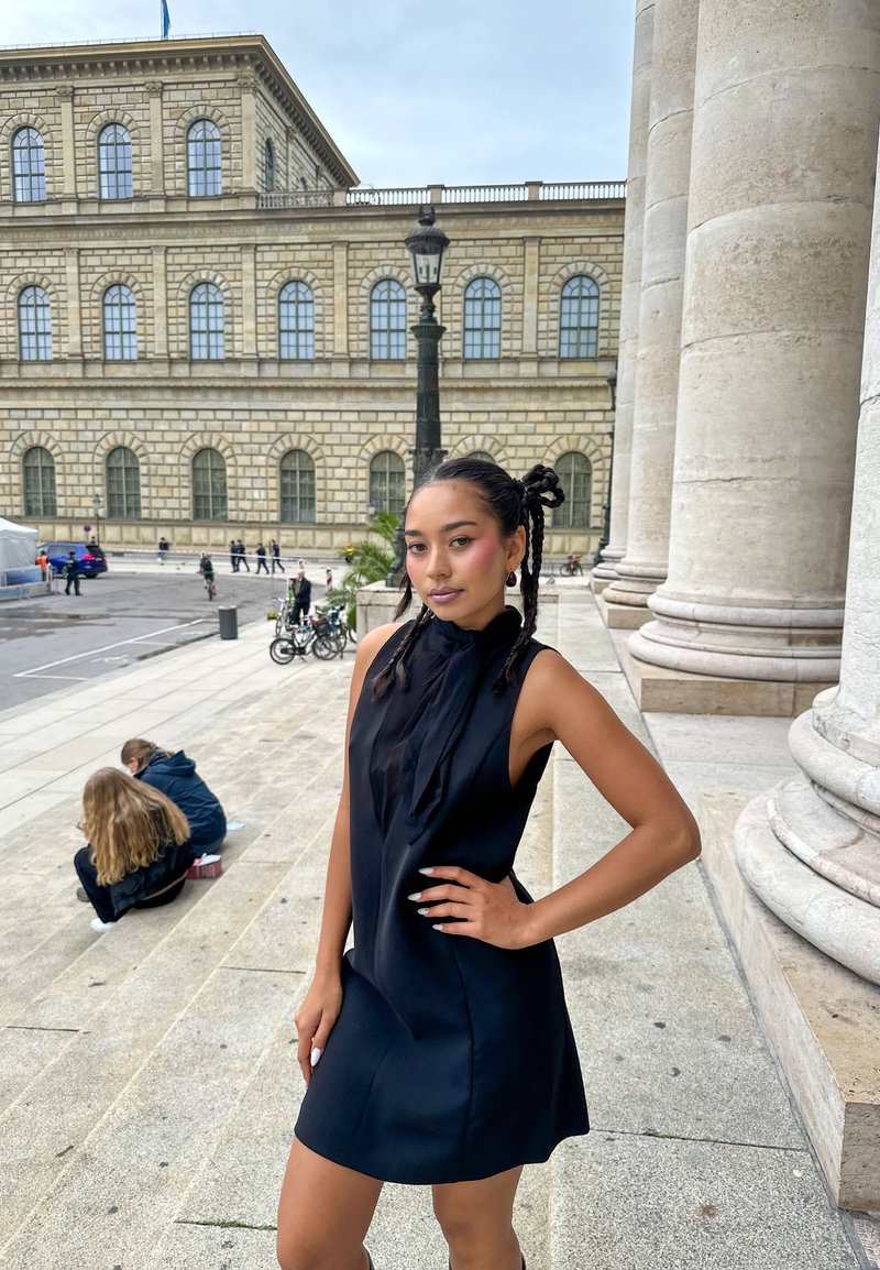 Giovane donna con capelli intrecciati in un vestito nero senza maniche che posa sui gradini di pietra di un edificio storico con grandi colonne e finestre ad arco.