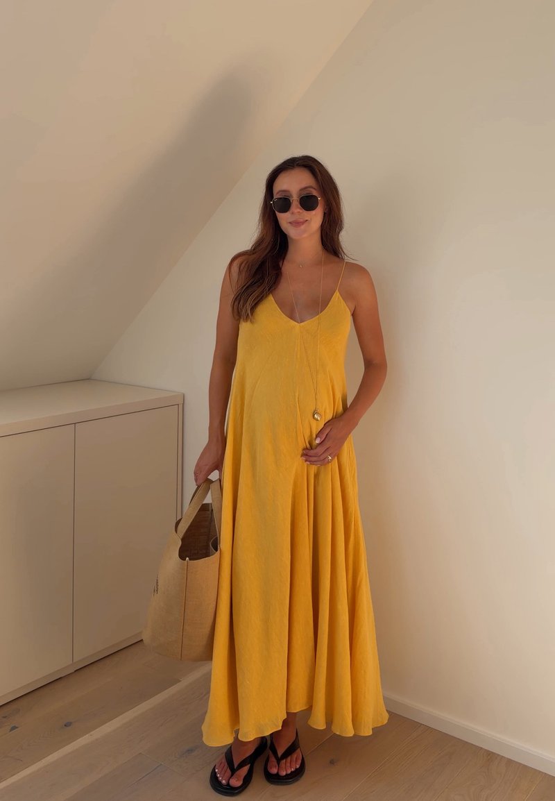 Femme portant une robe jaune sans manches, lunettes de soleil noires, tenant un sac fourre-tout beige, debout à l'intérieur contre un mur clair et un sol en bois.
