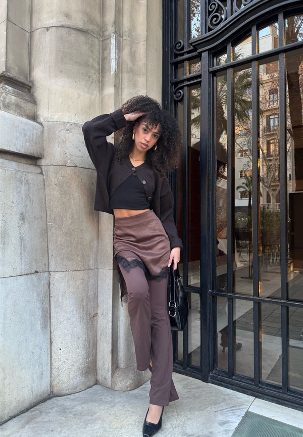 Sieviete ar cirtainiem matiem pozē pret akmens sienu, tērpusies melnā īsā topā, brūnā mežģīņu svārkā, brūnās biksēs un melnos apavos, turot melnu somu.