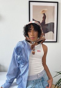 Blå randig skjorta med rullade tygdetaljer, vit linne med logotyp och mönstrad scarf. Bakgrunden har en inramad konstverks.