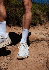 Ténis de desporto brancos com uma superfície texturizada, atacadores e uma aparência gastada, combinados com meias brancas até ao tornozelo, em um caminho de terra rochosa.