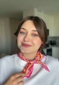 La sciarpa di seta presenta un motivo floreale con accenti rossi, arancioni e rosa, rifinita con un bordo rosa uniforme. La modella la indossa annodata al collo.