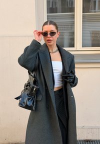 Νεαρή γυναίκα με μαλλιά χτενισμένα προς τα πίσω, φορώντας φαρδιά γκρι παλτό, μαύρα γάντια, γυαλιά ηλίου, ασημένιο κολιέ, κρατώντας μαύρη δερμάτινη τσάντα, στέκεται σε εξωτερικό χώρο.