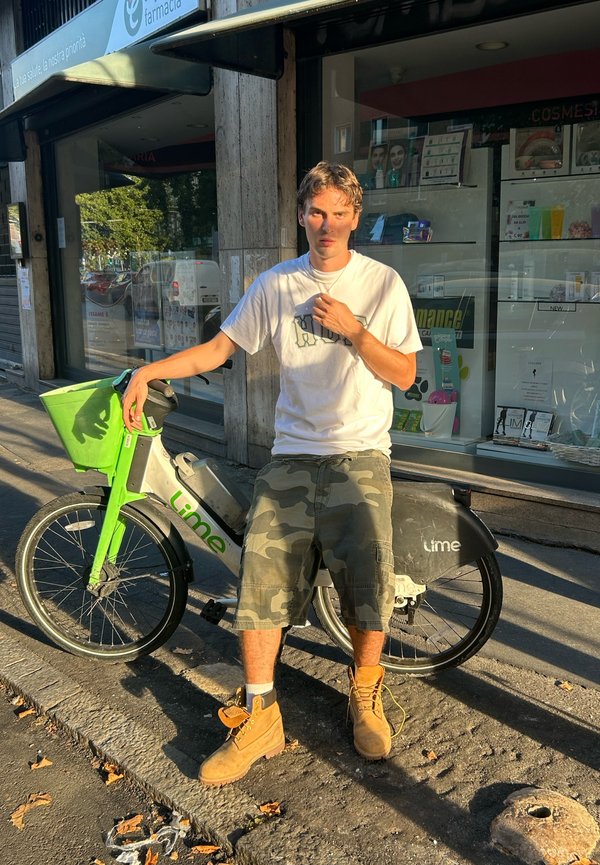 Lime elektriskais velosipēds ar zaļu priekšējo grozu un pelēku rāmi. Braucējs baltā T-kreklā un kamuflāžas šortos stāv blakus tam uz bruģētas ielas.