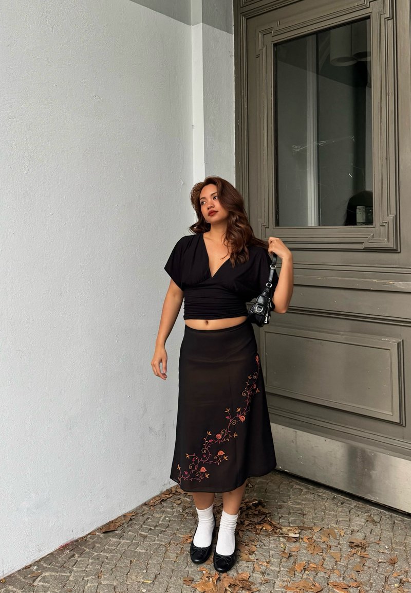 Femme aux cheveux ondulés se tient près d'une grande porte grise, portant un crop top noir, une jupe à fleurs noire, des chaussettes blanches et des ballerines noires, tenant un petit sac.