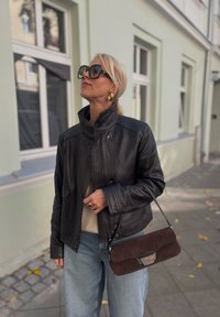 Črna usnjena jakna z visokim ovratnikom, v kombinaciji s svetlim bež puloverjem in širokimi svetlimi jeans hlačami. Rjava semiš torbica z kovinskimi detajli.