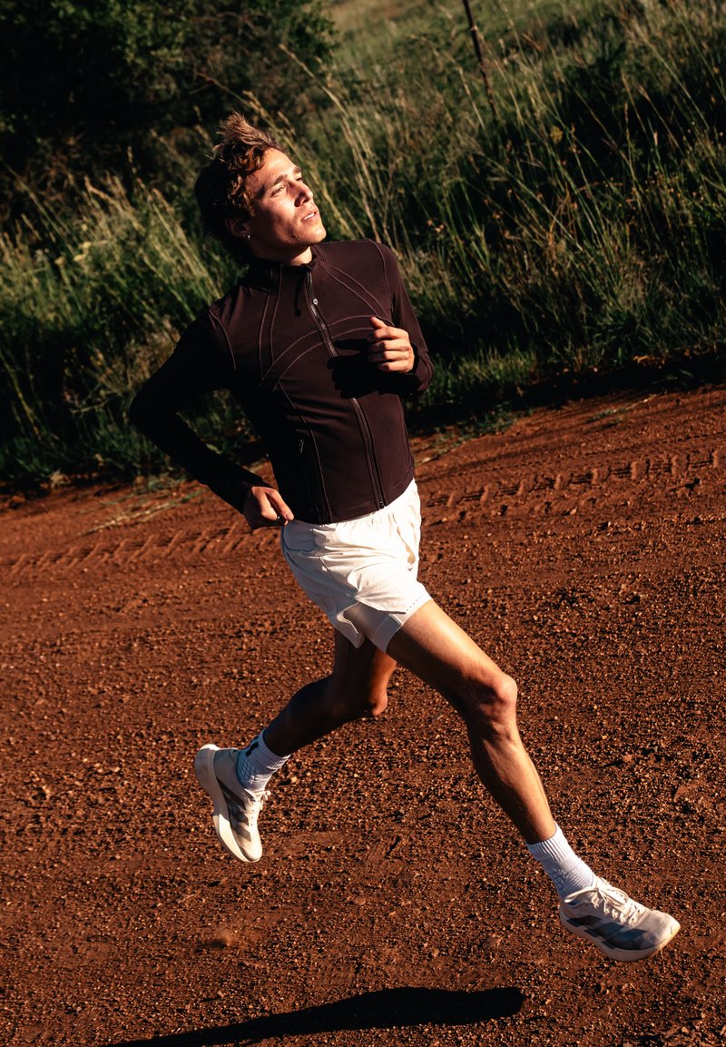 Coureur vêtu d'une veste noire, short blanc et baskets, courant sur un sentier de terre avec des hautes herbes en arrière-plan sous un soleil éclatant.
