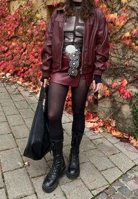 Nainen pukeutuneena viininpunaiseen nahkatakkiin ja hameeseen, musta sukkaisiin ja saappaisiin, kädessään musta laukku ja viininpunaiset aurinkolasit, seisoo syksyn lehtien lähellä.
