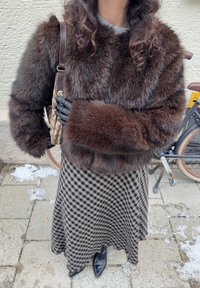 Braune Jacke aus Kunstpelz mit weiten Ärmeln, kombiniert mit einem Rock im schwarz-weiß karierten Muster. Schwarze Handschuhe und Accessoires sind sichtbar.