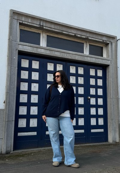 Mujer con cabello oscuro, gafas de sol, cárdigan azul marino, camisa blanca y jeans azul claro, de pie frente a una puerta azul con paneles cuadrados blancos.
