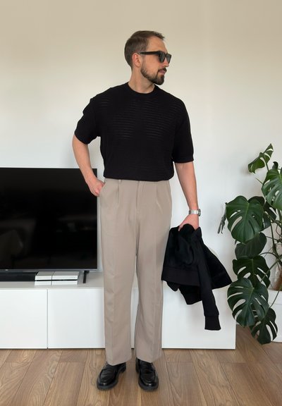 Suéter negro de punto de manga corta, pantalones beige con pliegues, zapatos negros. La persona sostiene una chaqueta negra, posando junto a un televisor.
