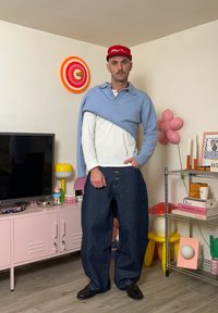 Modell trägt einen hellblauen Wickelpullover, weißes Hemd und dunkelblaue, weite Jeans, dazu schwarze Schuhe. Im Hintergrund bunte Dekoration und Möbel.