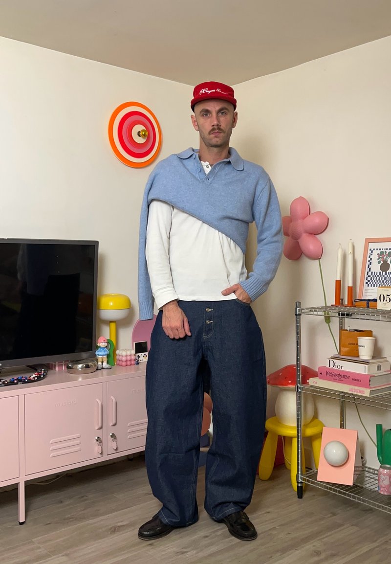 Modello che indossa un maglione avvolgente azzurro chiaro, camicia bianca e jeans scuri larghi, con scarpe nere. Lo sfondo presenta decorazioni e mobili colorati.