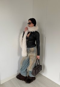 Veste en cuir noire, jean bleu délavé oversize, écharpe blanche et moelleuse, sac à main marron, et bottes marron fourrées, le tout sur un fond uni.