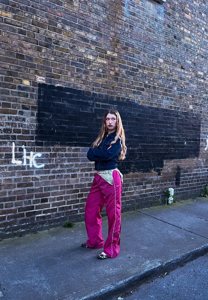 Jeune femme aux longs cheveux se tient sur le trottoir près d'un mur en briques avec des graffitis, portant une veste en jean et un pantalon rose vif, les bras croisés.