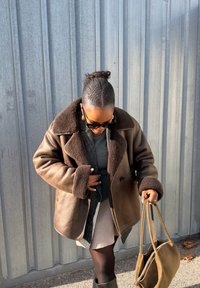 Femme aux cheveux tressés portant des lunettes de soleil foncées, un manteau en peau retournée marron, une jupe courte, des collants noirs et tenant un sac à main en daim marron en extérieur.