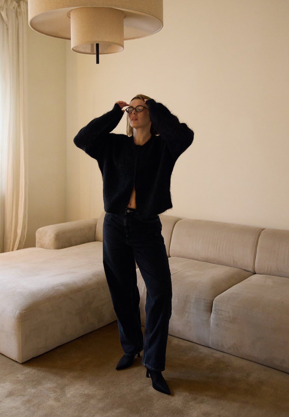 Melna, pūkaina, īsā džemperīša ar plašām piedurknēm kompozīcija, kombinācijā ar tumšām džinsām un melniem, punktējošiem papēžiem. Gaiši bēša dīvāns un piekaramā gaisma fonā.