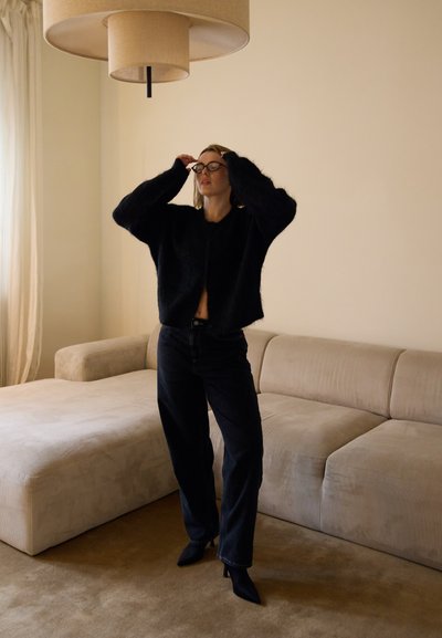 Suéter negro y peludo, recortado, con mangas anchas, combinado con jeans oscuros y tacones negros de punta. Sofá beige claro y lámpara colgante al fondo.