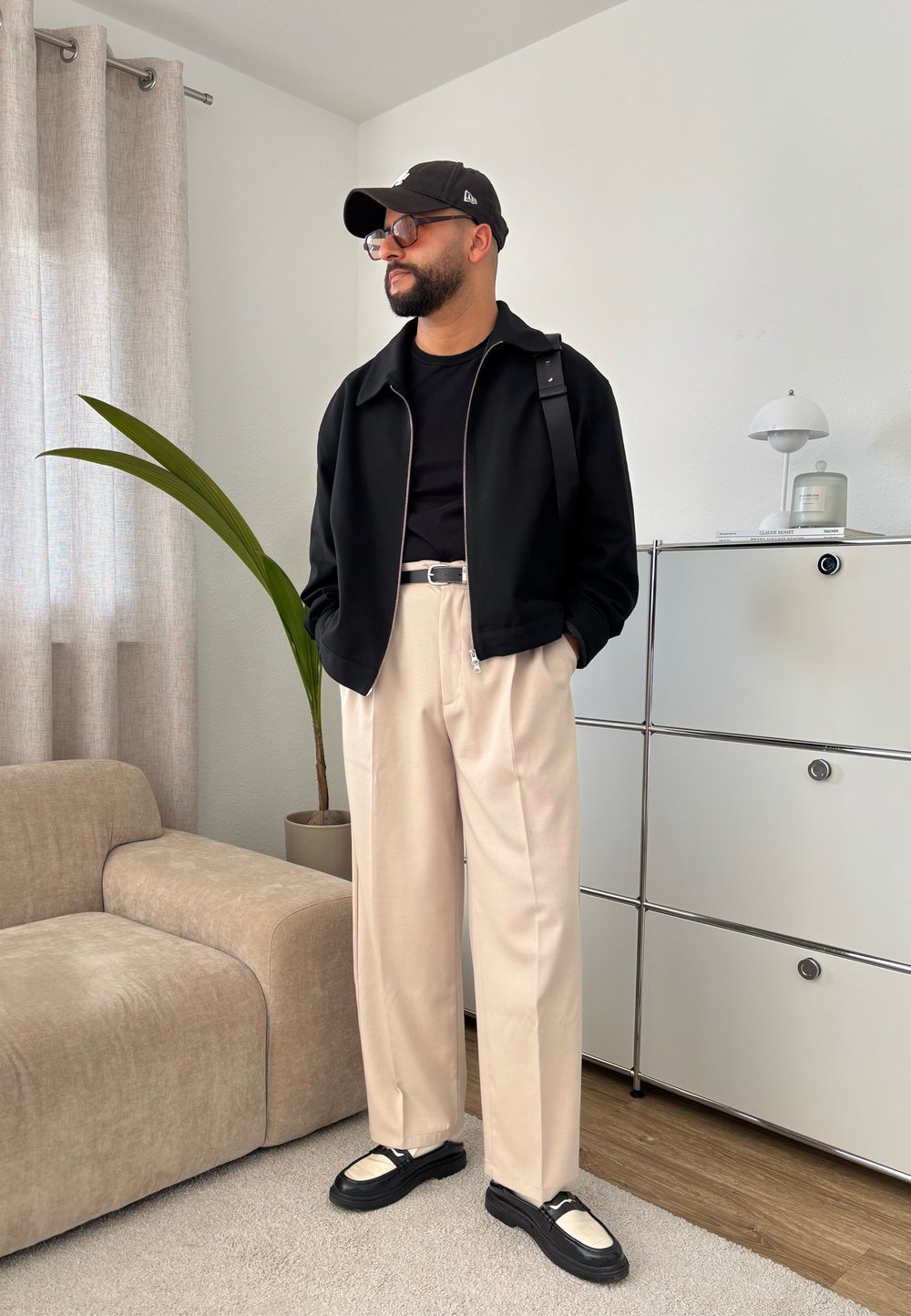 Crni zip-up dukserica preko crne majice, bež hlače i crne tenisice s bijelim naglascima. Unutarnje okruženje s biljkom i namještajem.